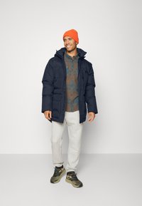 Jachetă puffer de culoare albastru închis cu glugă, cu două buzunare frontale, purtată peste o cămașă în carouri maro și teal, asortată cu pantaloni de trening gri și adidași verzi.