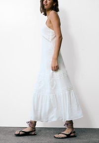 Robe maxi blanche avec fines bretelles, détails en crochet, ourlet à volants et accents en dentelle. Sandales plates noires avec brides tressées.