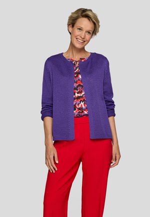 Smiling woman wearing a purple knitted cardigan over a patterned top and bright red pants, standing against a plain light gray background.
