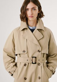 Beige trenchcoat med dubbelknappning, stor krage och ett tygbälte. Inkluderar knappdetaljer och sidofickor.