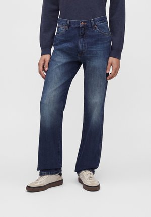 Man draagt donkerblauwe rechtvallende jeans, beige sneakers en een marineblauw shirt met lange mouwen, staat met ontspannen handen langs zijn zij.