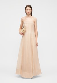 Frau trägt ein ärmelloses, bodenlanges beiges Kleid und hält eine kleine passende Handtasche vor einem schlichten hellen Hintergrund.