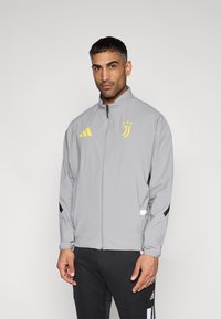 Ανοιχτό γκρι ζακέτα με φερμουάρ, που διαθέτει κίτρινο λογότυπο Adidas, έμβλημα της Γιουβέντους και μαύρες λεπτομέρειες στα μανίκια· κατασκευασμένη από ελαφρύ υλικό.