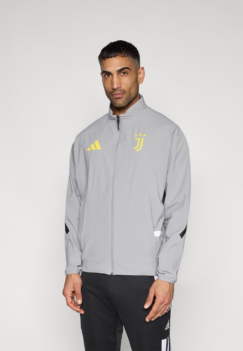 Ανοιχτό γκρι ζακέτα με φερμουάρ, που διαθέτει κίτρινο λογότυπο Adidas, έμβλημα της Γιουβέντους και μαύρες λεπτομέρειες στα μανίκια· κατασκευασμένη από ελαφρύ υλικό.