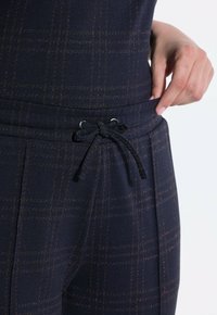 Ruka upravující pás nohavic modro-černý kostkovaný vzor s utaženou šňůrkou a viditelnou přední kapsou.