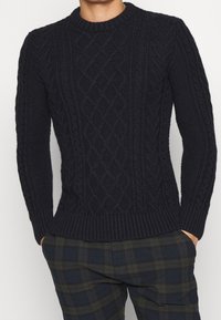 Pull en maille torsadée bleu marine à col rond, poignets et ourlet côtelés ; des motifs texturés créent un effet de superposition sur un fond lisse.