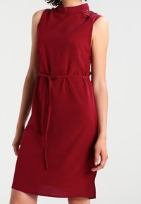 Robe sans manches bordeaux avec un col montant, des accents en dentelle sur les épaules et une ceinture en tissu à la taille, présentant une texture lisse et fluide.