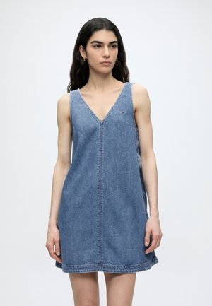 Kvinde med langt mørkt hår iført ærmeløs blå denimkjole med V-hals og lille logo på brystet, stående mod en ensfarvet baggrund.