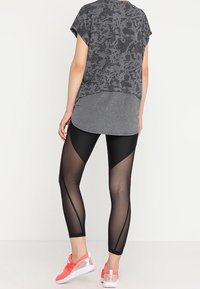 Grå sporttröja med ett mönstrat design, kombinerad med svarta leggings med genomskinliga paneler och geometriska accenter. Rosa sneakers fulländar looken.