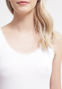 Biały top na ramiączkach z falbaniastą koronką przy dekolcie i armhole'ach, wykonany z miękkiego, lekkiego materiału.