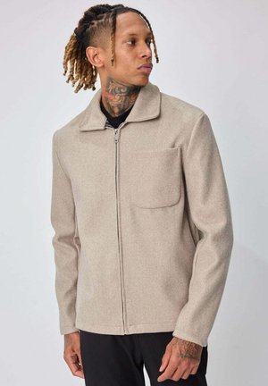 Homme aux cheveux tressés et tatouage au cou, portant une veste beige zippée avec une poche poitrine, regardant sur le côté sur un fond uni.