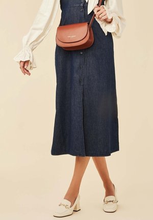 Kobieta ubrana w ciemną jeansową spódnicę zapinaną na guziki, białą bluzkę z falbankami, białe mokasyny na obcasie oraz małą brązową torebkę na ramię.