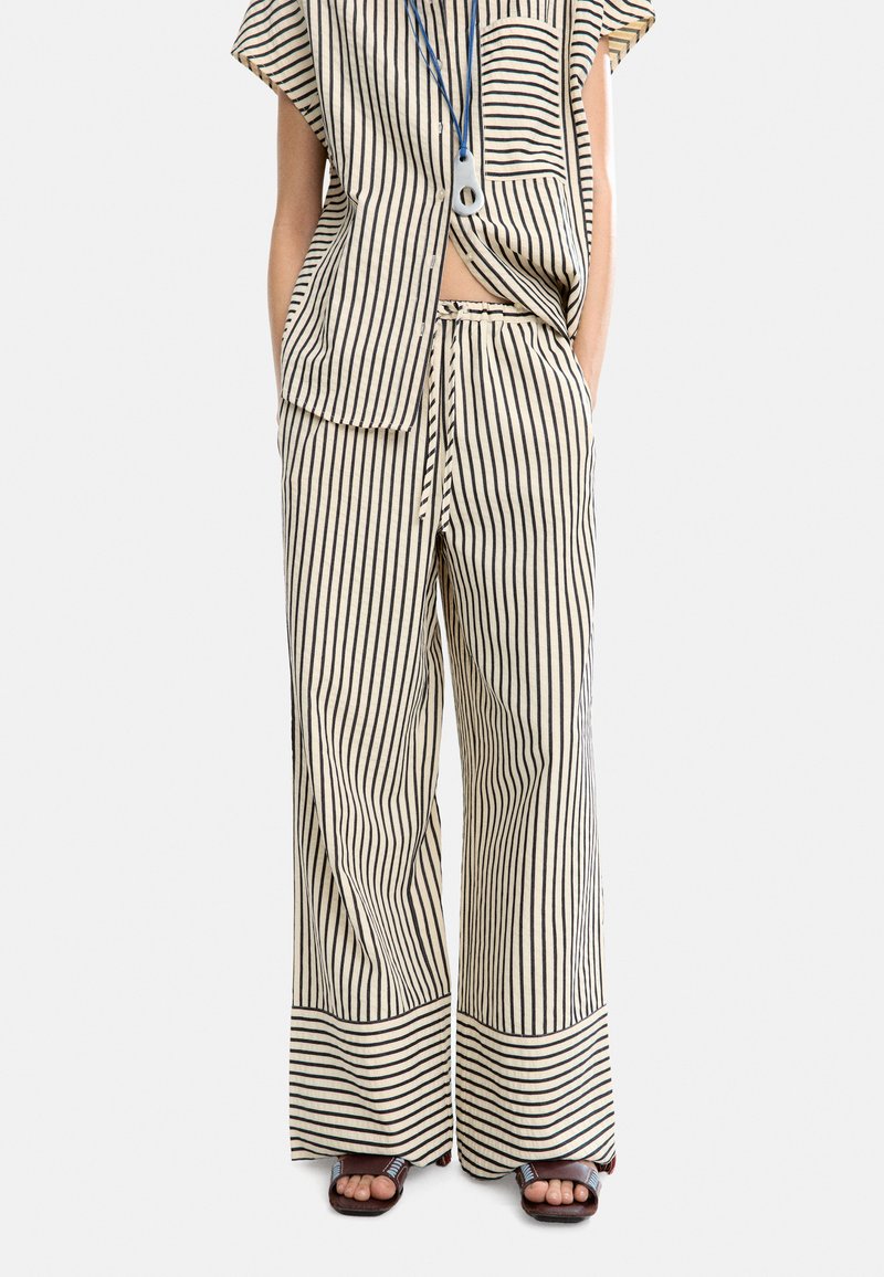 Set coordinato in cotone a righe, composto da una maglietta a maniche corte e pantaloni a gamba larga, nei colori crema e nero. Dettagli notevoli includono tasche laterali e rifiniture ai bordi.