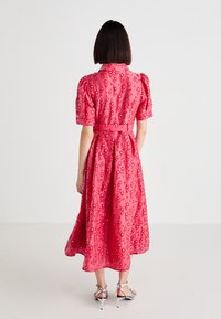 Mujer vistiendo un vestido rosa hasta la rodilla con bordado floral y mangas abullonadas, de pie de espaldas sobre un fondo blanco liso.