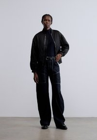 Crna bomber jakna preko crnog rolka; tamne traperice visokog struka s vidljivim šavovima. Stoji nasuprot svijetle pozadine.