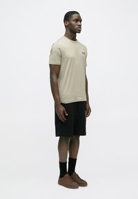 Helebeežist puuvillane T-särk väikes logo rinnal, mustade lühikeste pükstega, millel on külgedel tekst, ja pruunid nööritavad kingad.