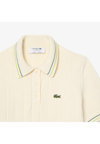 Polo di colore crema con texture a coste, apertura con tre bottoni e colletto a righe verdi e gialle. Presenta il logo ricamato Lacoste.