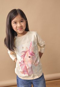 Chemise à manches longues crème avec un graphique de licorne, crinière rose, détails scintillants et étoiles ; associée à un jean bleu.