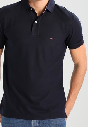 Mann trägt ein eng anliegendes dunkelblaues Polohemd mit kurzen Ärmeln und einem kleinen rot-weiß-blauen Logo auf der linken Brust.