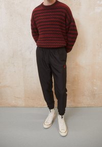 Red and black striped sweater, relaxed fit. Black tapered pants with elastic waistband and small red logo. White high-top sneakers.
