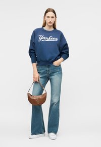 Frau trägt einen blauen Yankees-Sweatshirt, ausgestellte Jeans, weiße Turnschuhe, hält eine braune Lederhandtasche und steht vor einem schlichten Hintergrund.