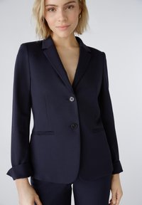 Ein Marineblazer aus glattem Stoff mit einem V-Ausschnitt, zwei Knöpfen und aufgesetzten Taschen. Die umschlagbaren Ärmel verleihen dem maßgeschneiderten Design eine besondere Note.