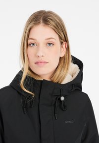 Veste noire imperméable avec un intérieur en beige doux, capuche réglable et cordons de serrage. Présente un logo près de la fermeture éclair.