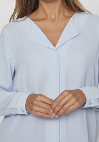 Blouse bleu clair avec col cranté, boutons sur le devant et manches longues. Le tissu semble lisse avec un léger éclat et boutons aux poignets.