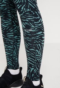 Tight-fitting leggings med ett mönster av teal zebra-ränder på en svart bakgrund. Visad logotyp i vitt på vänster ben.