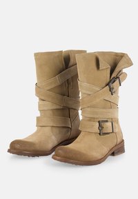 Botas de ante de media caña en color marrón claro con correas cruzadas y detalles de hebilla metálica. Punta redondeada, superficie texturizada y tacón de madera apilada.