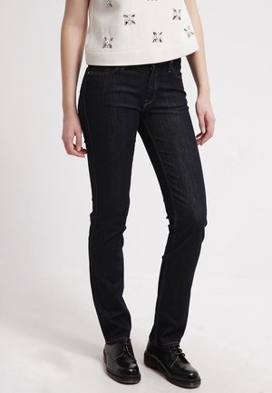 Vrouw draagt strakke donkerblauwe jeans, een witte mouwloze top met versieringen en zwarte veterschoenen, staand tegen een effen achtergrond.