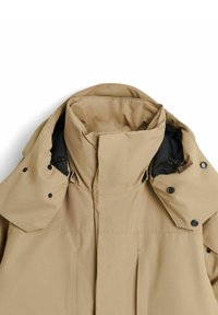 Beige vattentät jacka med hög krage, svarta detaljer, tryckknappar och en texturerad tyg. Har en kortad design med veck.