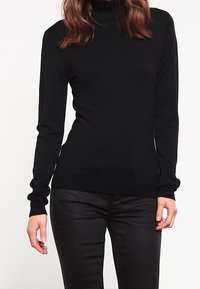 Pull à col roulé noir en tissu doux, coupe ajustée, manches longues et ourlet côtelé. Associé à un pantalon noir pour un look monochrome.