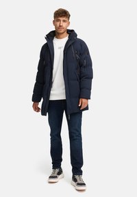 Marinblå puffjacka med huva, dragkedja framtill, dragkedjefickor och quiltad textur. Bärs över en vit topp och mörka jeans.