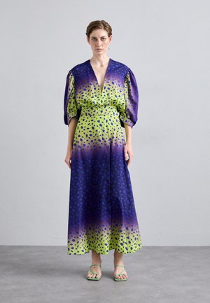Lila und grün verlaufendes Kleid mit Puffärmeln, tiefem V-Ausschnitt und gepunktetem Muster; kombiniert mit grünen Riemchensandalen.