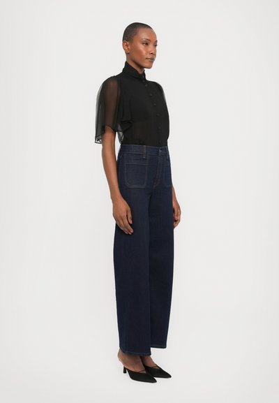 Black sheer blouse with short, flowing sleeves, paired with high-waisted dark blue denim wide-leg trousers. Matte black heels complete the look.