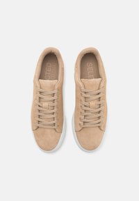 Beżowe zamszowe trampki z sześcioma oczkami i dopasowanymi sznurówkami, biała gumowa podeszwa oraz wytłoczone logo na wkładce. Gładka tekstura i prosty design.