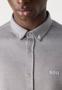 Šedá polo košeľa s vzorom, so zapínaním na gombíky a bielym logom "BOSS" na ľavej strane.