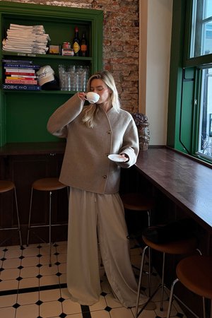 Veste en mélange de laine beige avec un col haut et des boutons, accompagnée de pantalons légers et fluides. Tasse et soucoupe blanches à la main, se tenant au bar.