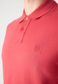 Polo rouge avec deux boutons et un petit logo brodé "BOSS" sur la poitrine gauche, porté par une personne.