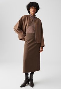Giacca marrone con zip realizzata in materiale morbido, abbinata a un top marrone aderente e a una gonna marrone fino al ginocchio, accentuata da stivali neri alla caviglia.