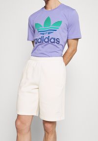 Lila bomull t-shirt med gröna och blå randiga Adidas-logotyp, ihopparad med vita fleece-shorts med elastiskt midjeband och sidfickor.