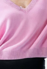 Maglione rosa a maglia con scollatura a V e orlo a coste. Presenta una texture morbida e un singolo bottone decorativo sulla scollatura.