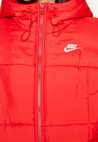 Röd vadderad jacka med huva, utrustad med en främre dragkedja och snörning, samt en vit Nike-logotyp på vänster sida. Slät nylontextur.