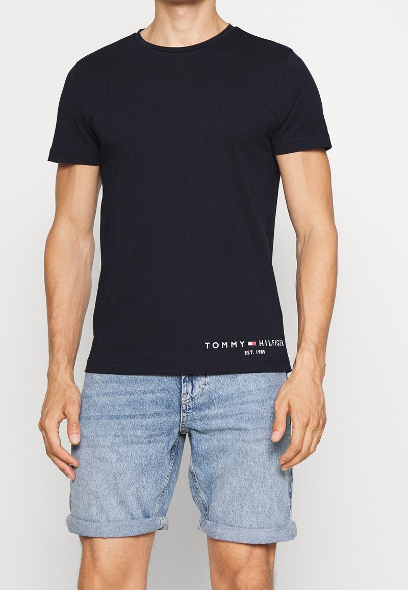 Mężczyzna ubrany w czarną koszulkę Tommy Hilfiger z logo na dolnej krawędzi oraz jasnoniebieskie, podwinięte dżinsowe szorty, stojący na tle białym.