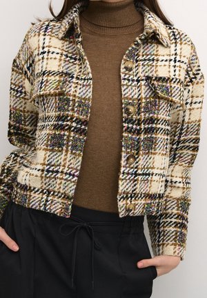 Vrouw die een beige en zwart geruit jasje met knopen draagt over een bruine coltrui en zwarte broek met trekkoord, met haar handen in haar zakken.