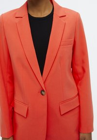 Blazer de color coral en tejido ligero con un corte entallado, cierre de un solo botón y dos bolsillos frontales. Presenta solapas con muescas y hombros estructurados.