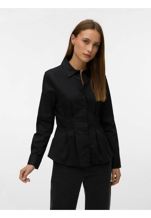 Femme aux cheveux bruns lisses portant une chemise noire cintrée à boutons avec des plis et un pantalon noir sur un fond blanc uni.
