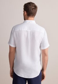 Chemise en lin blanc à manches courtes avec un ourlet arrondi, un pli dans le dos et un col boutonné, présentant un tissu texturé et des poignets retroussés.