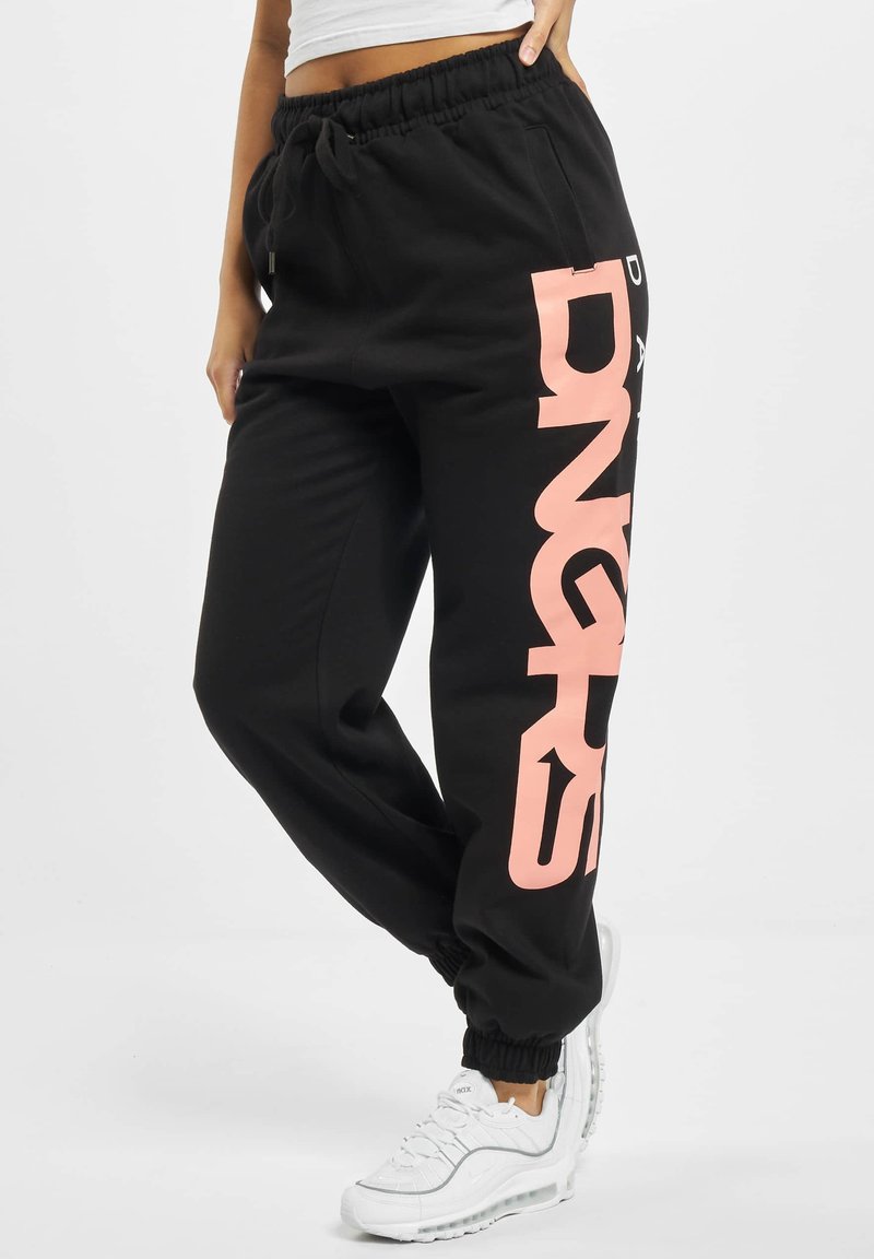 Black sweatpants feature a large pink text graphic on the left leg, elastic waistband, and cuffs. Paired with white athletic shoes.